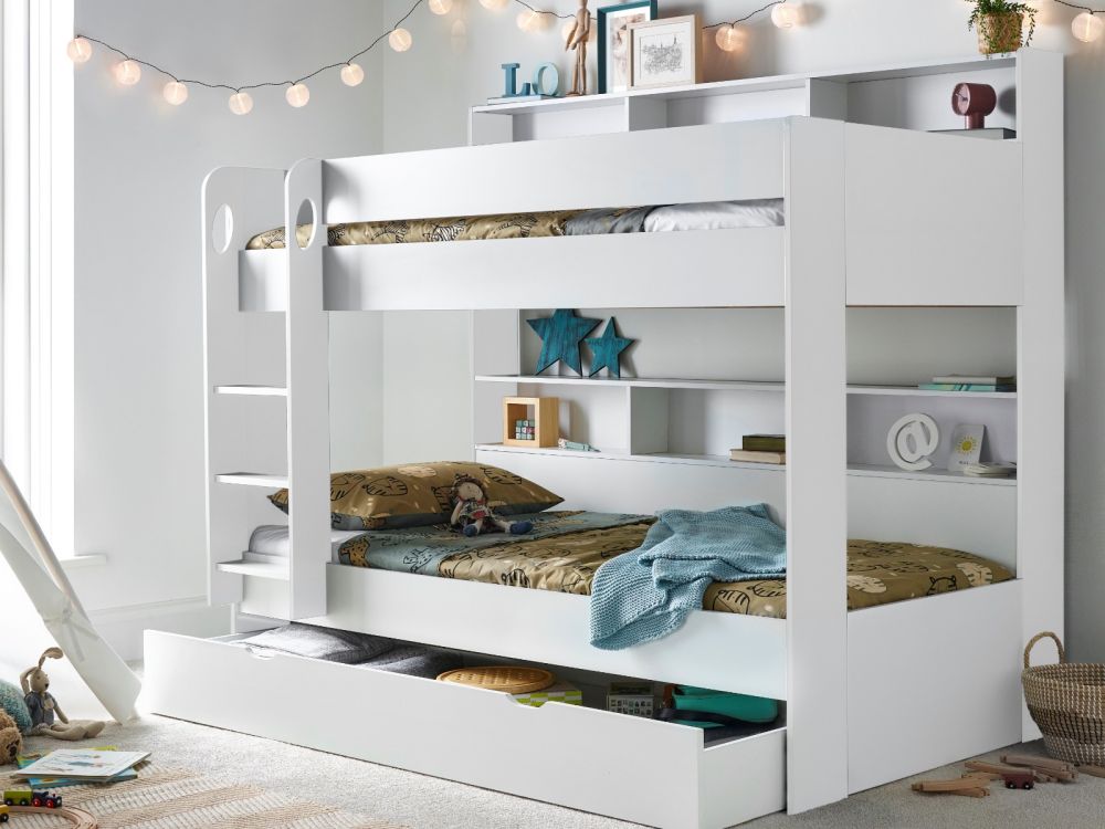 White wooden bunk bed with a storage drawer and built-in shelves, with the bed dressed with multicolored bedding and the room lit with string lights.